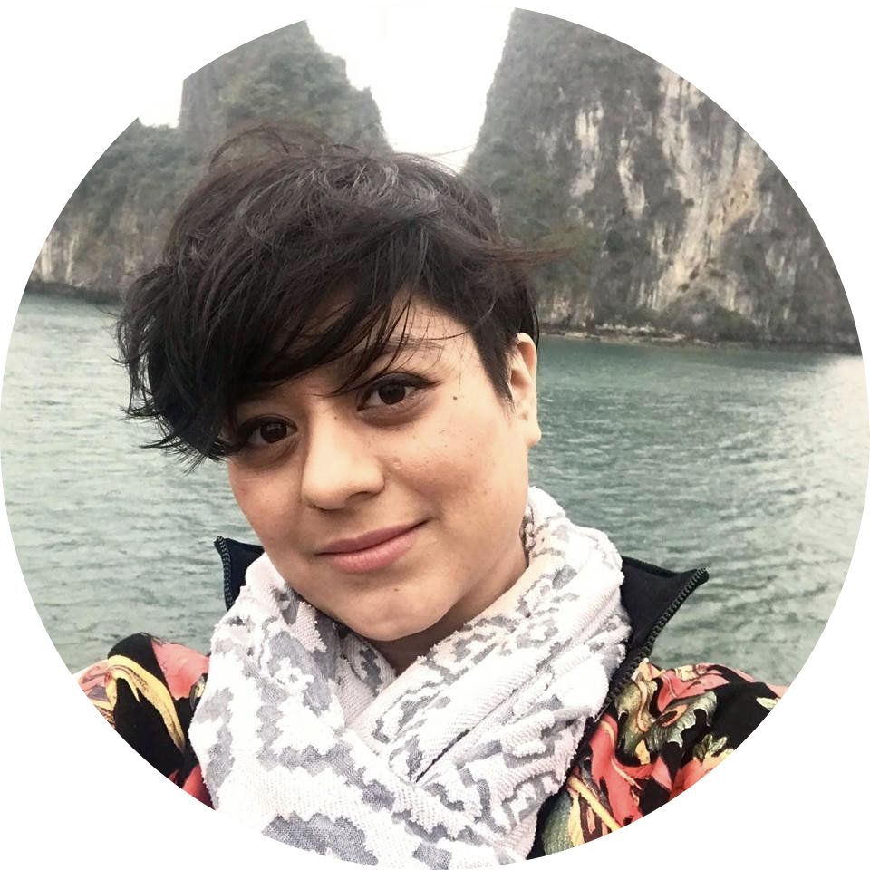 Person with short dark hair wearing a scarf and jacket, posing outdoors by rocky cliffs and water.
