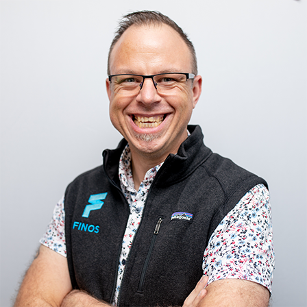 Smiling man with glasses wearing a black FINOS vest over a floral shirt, arms crossed.