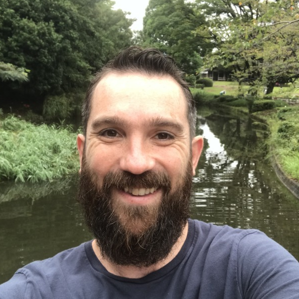 Bearded man smiling outdoors by a calm river with green trees and grass in the background.