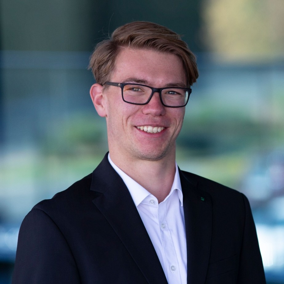 Smiling young man with glasses in a black suit and white shirt posing against blurred background.