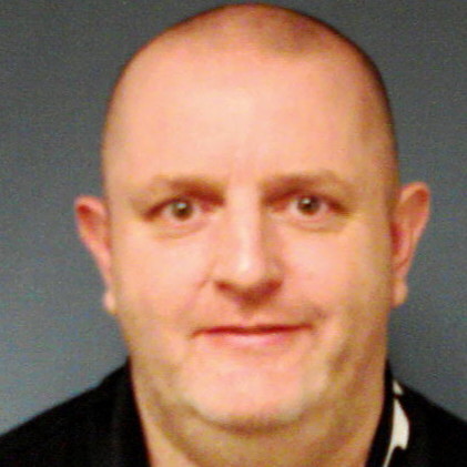 Smiling bald man with light skin and brown eyes wearing a black shirt against a blue background.
