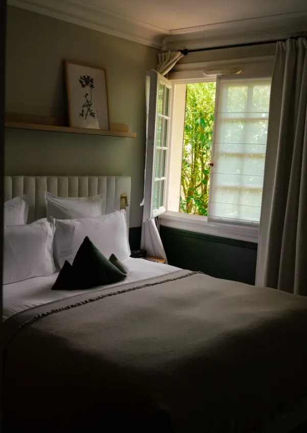 Camera da letto con letto matrimoniale, cuscini bianchi, cuscini scuri, finestra aperta che lascia entrare la luce e vista sugli alberi verdi.