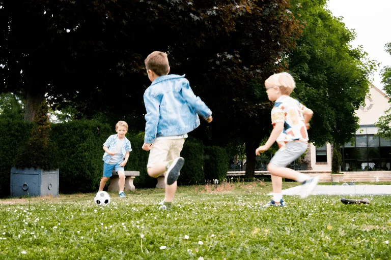 Three children are playing soccer in a garden with trees and a house in the background.
