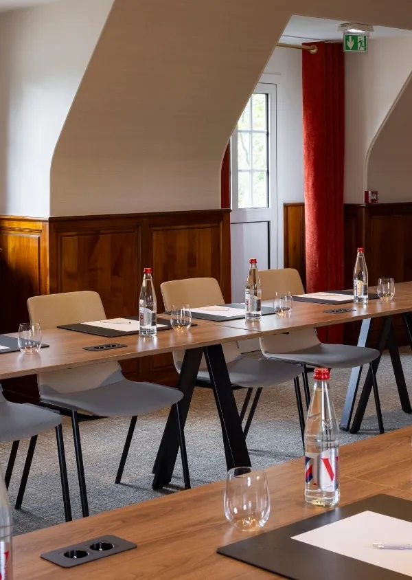 Salle de réunion avec table en bois, chaises beige, bouteilles d'eau et verres disposés, décor en bois et rideaux rouges.