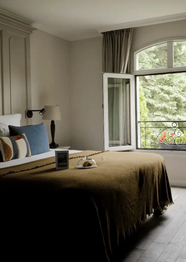 Schlafzimmer mit Doppelbett, bezogen mit einer braunen Decke, bunten Kissen, Nachttischlampe und offenem Fenster mit Blick auf grüne Bäume.
