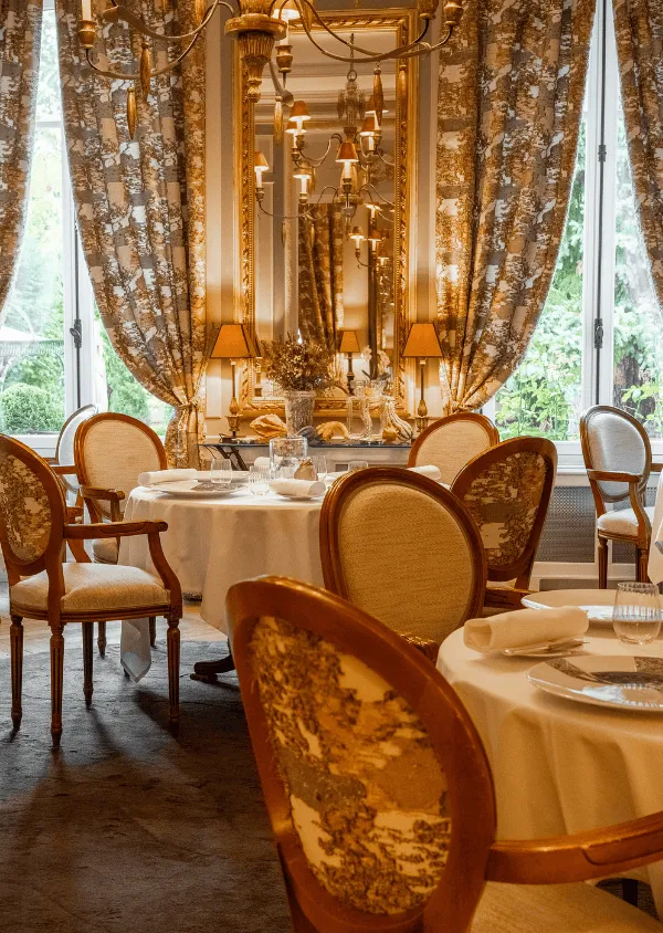 Elegantes Esszimmer mit Holz- und Stoffstühlen, runden Tischdecken und großen Fenstern, die mit floralen Vorhängen geschmückt sind.