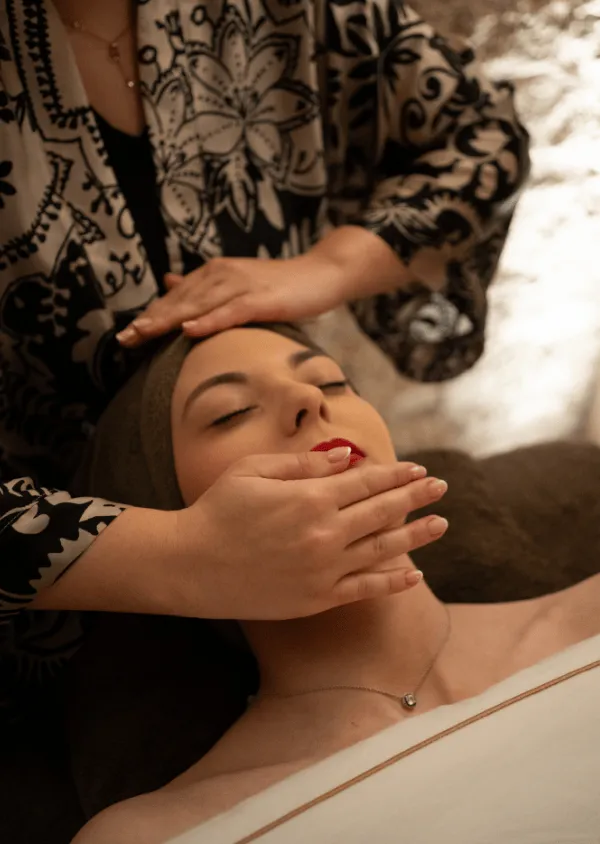 Uma mulher recebe um tratamento facial com as mãos de uma esteticista com os olhos fechados e um lençol branco sobre o corpo.