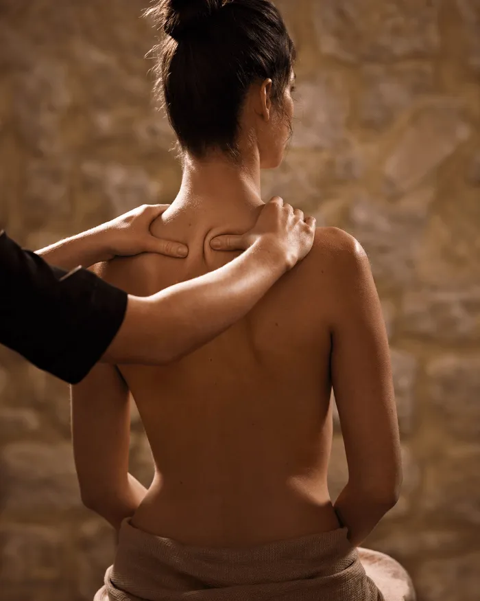 Eine von hinten gesehene Frau erhält eine Schultermassage in einer warmen Umgebung mit einer verschwommenen Steinmauer im Hintergrund.
