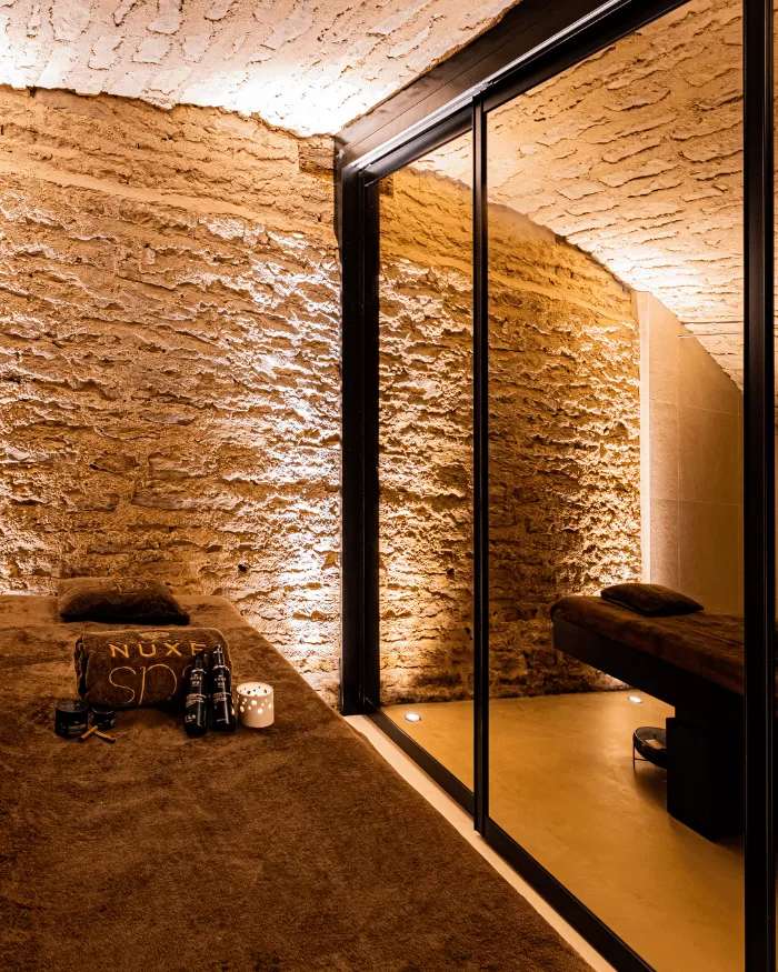 Warmer Spa-Raum mit beleuchteten Steinwänden, zwei braunen Massageliegen und Nuxe-Pflegeprodukten.
