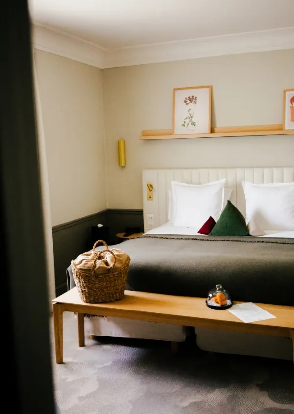 Modern hotel room with a double bed, decorative cushions, a wooden bench with a basket and a plate of food under a bell.