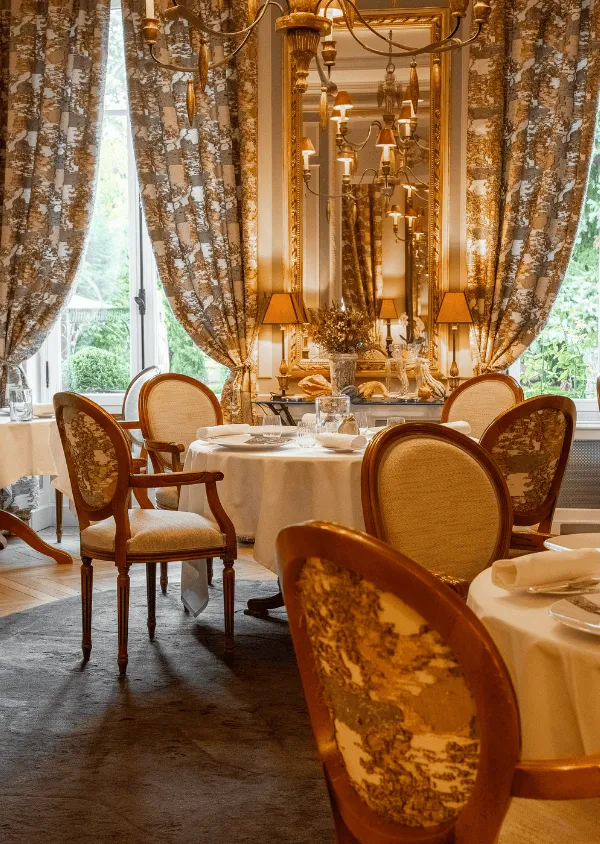 Stilvolles Esszimmer mit gedeckten runden Tischen, Holzstühlen mit Blumenmuster und großen Fenstern mit passenden Vorhängen.