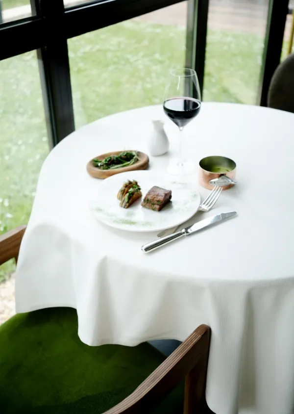 Tavolo rotondo con tovaglia bianca vicino a una finestra, pronto per un pasto con un bicchiere di vino rosso, un piatto con carne e verdure, posate e piccoli accessori.