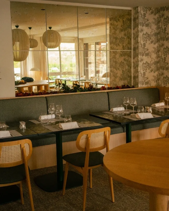 Sala da pranzo di un ristorante con panca grigia, tavoli apparecchiati con bicchieri da vino e tovaglioli, sedie in legno chiaro e un grande specchio sul muro.