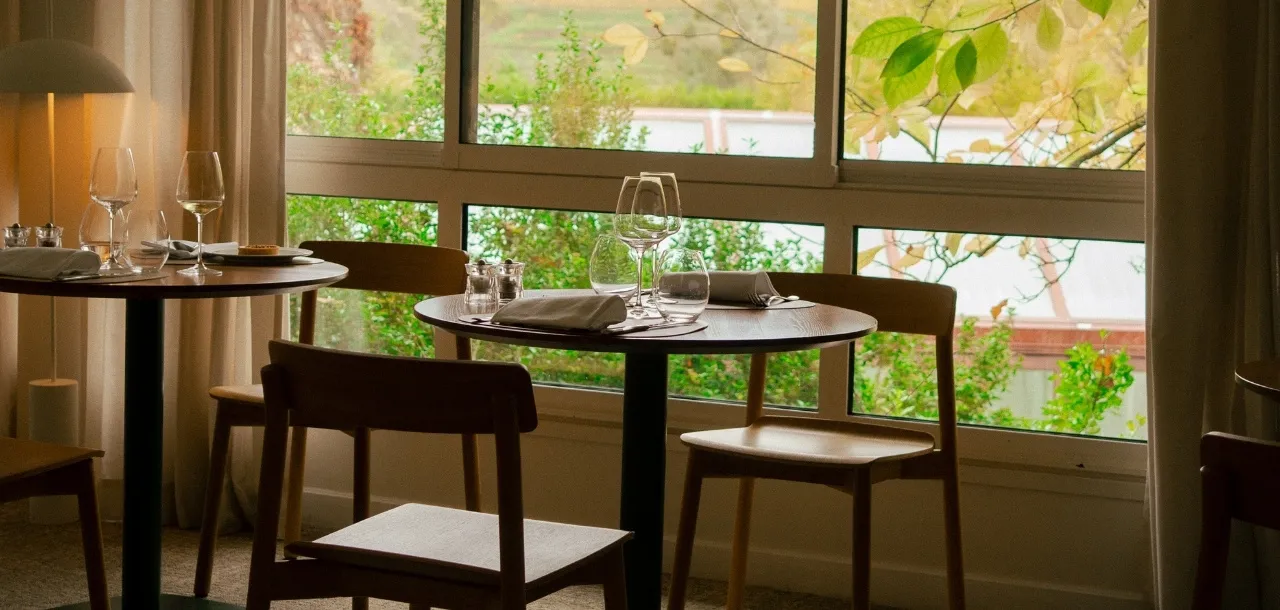 Una tavola rotonda apparecchiata con bicchieri da vino e tovaglioli in una luminosa sala da pranzo con una grande finestra che si affaccia su un giardino verde.