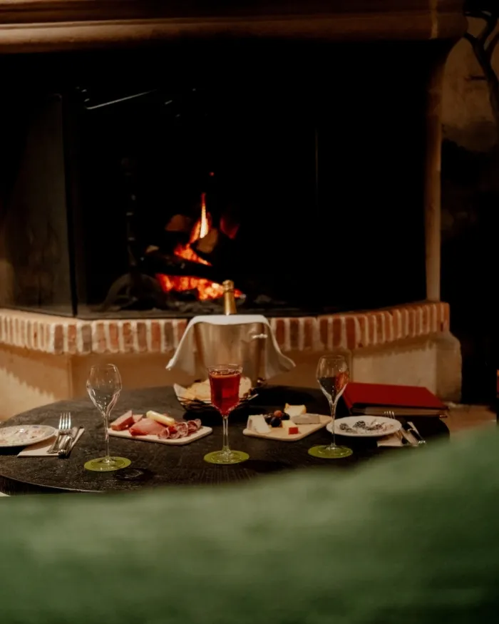 Tavola apparecchiata con un bicchiere di vino, taglieri di salumi e formaggio davanti a un camino acceso in una calda atmosfera.
