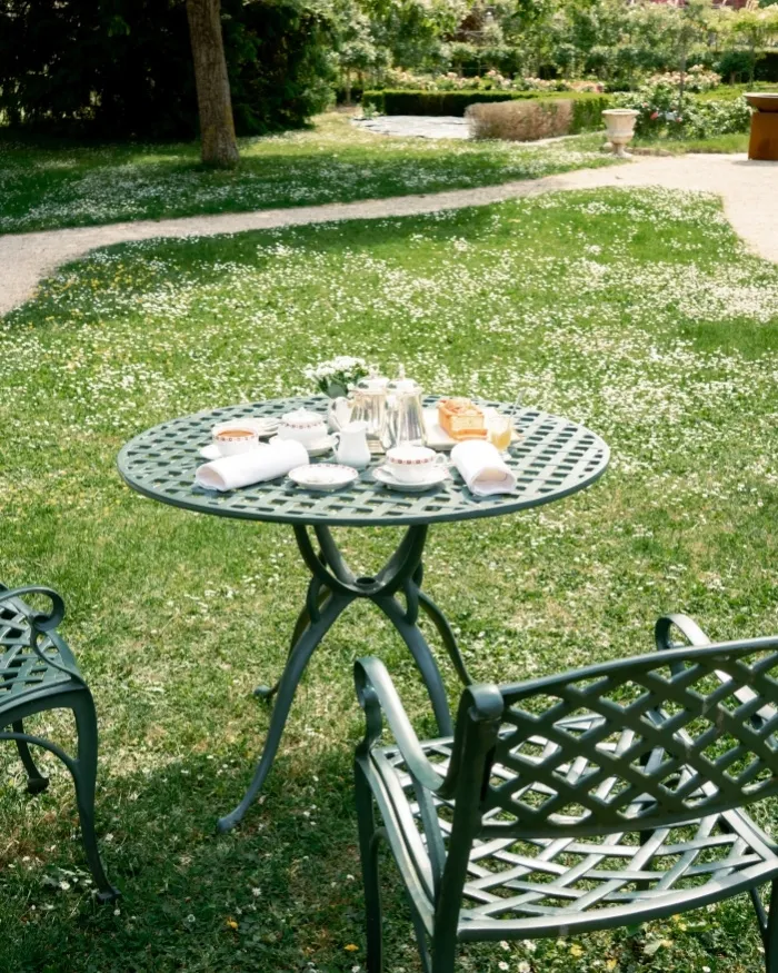 Tavolo rotondo in metallo con piatti e tovaglioli posto su un prato verde con fiori bianchi, circondato da sedie da giardino.