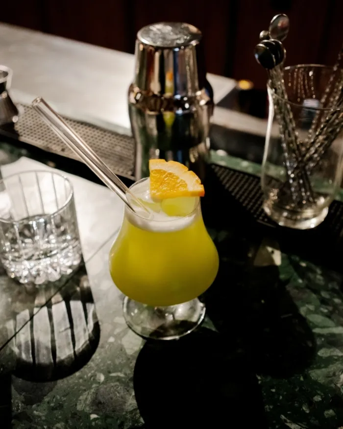 Cocktail giallo in un bicchiere con stelo, sormontato da una fetta d'arancia e una cannuccia trasparente su un bancone di marmo con uno shaker e cucchiai da bar sullo sfondo.