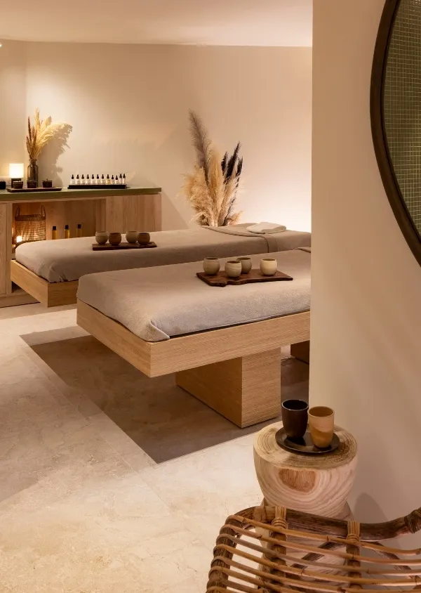 Salle de spa avec deux tables de massage en bois clair, décorée avec des objets en céramique et des plantes séchées dans un éclairage doux et chaleureux.