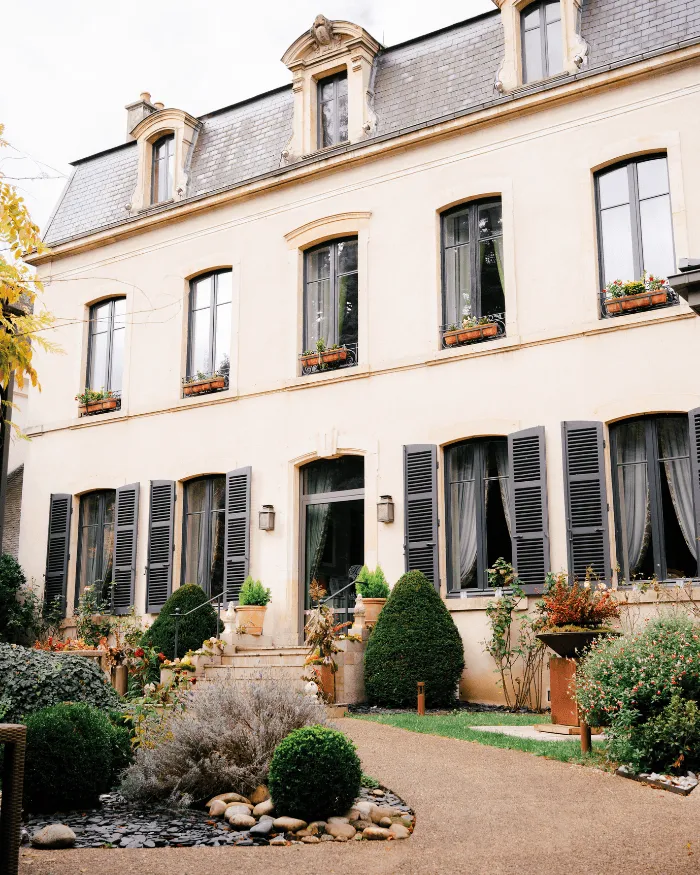 Fassade eines eleganten Hauses im klassischen Stil mit grauen Fensterläden, Blumentöpfen auf den Fensterbänken und einem gepflegten Garten mit verschiedenen Sträuchern und einem Kiesweg.