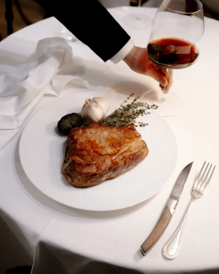 Ein saftiges Steak auf einem weißen Teller mit einer Knoblauchzehe, einem Trüffel und Thymianzweigen, neben einem Messer und einer Gabel auf einem weißen Tisch, eine Hand hält ein Glas Rotwein.