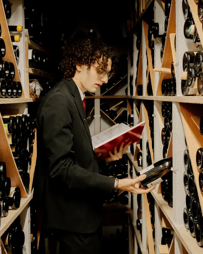 Ein Mann mit lockigem Haar und Brille betrachtet eine Flasche Wein in einem Keller voller Flaschenreihen.