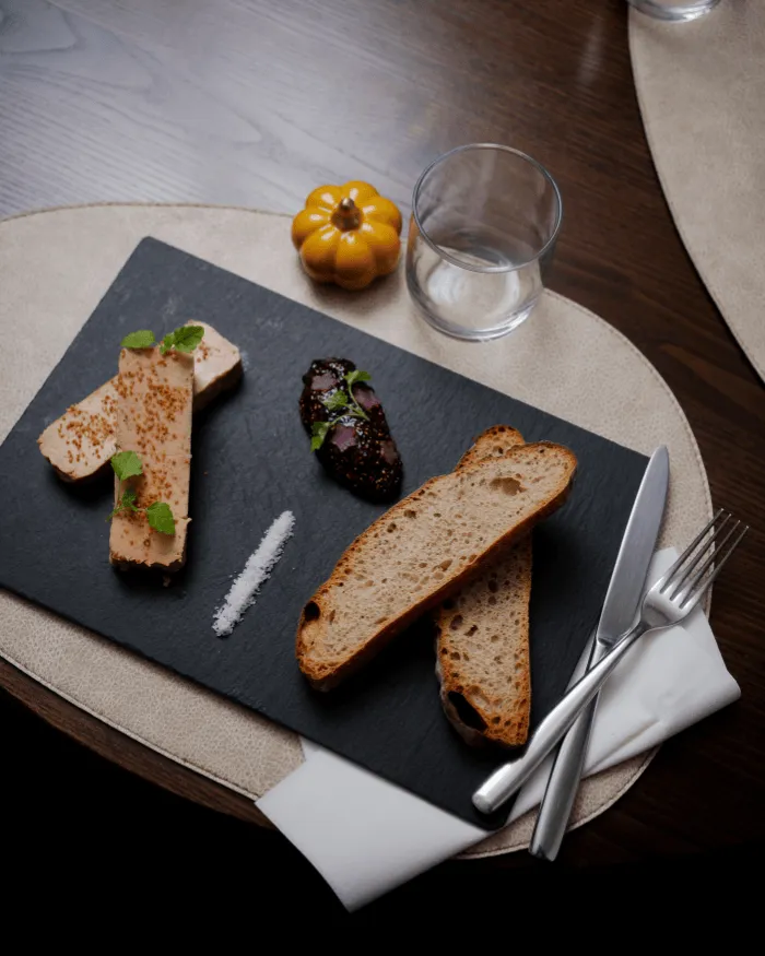 Schieferbrett mit zwei Scheiben Foie Gras, einem Löffel Marmelade oder Chutney, einer Linie Salz und zwei Scheiben Brot, serviert mit einem Messer und einer Gabel auf einer beigen Tischdecke.