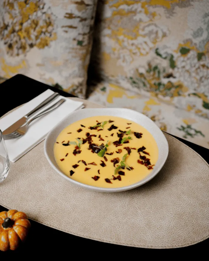 Schüssel mit cremegelber Suppe, garniert mit Kräuterblättern und knusprigen Stücken, auf einen beigen Teppich gelegt, mit Besteck und einem kleinen Kürbis daneben.