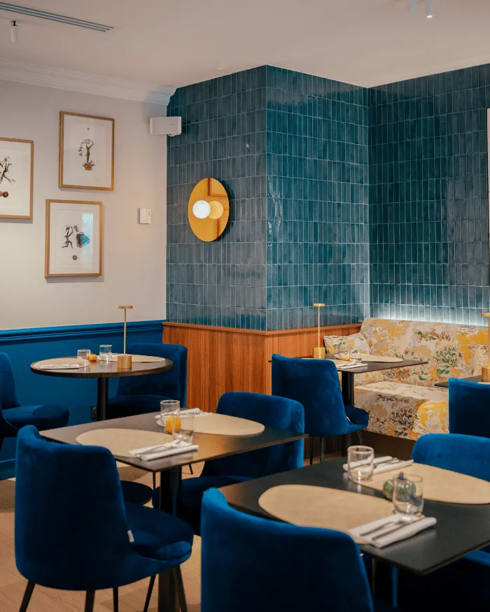 Modernes Café-Interieur mit marineblauen Stühlen, schwarzen und beigen Tischen, einer blau gefliesten Wand und einer gelben Blumenbank.