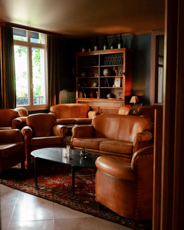 Warmes Wohnzimmer mit braunen Ledersofas und Sesseln rund um einen schwarzen Couchtisch, einem rot gemusterten Teppich und einem Bücherregal aus Holz vor einer dunklen Wand.