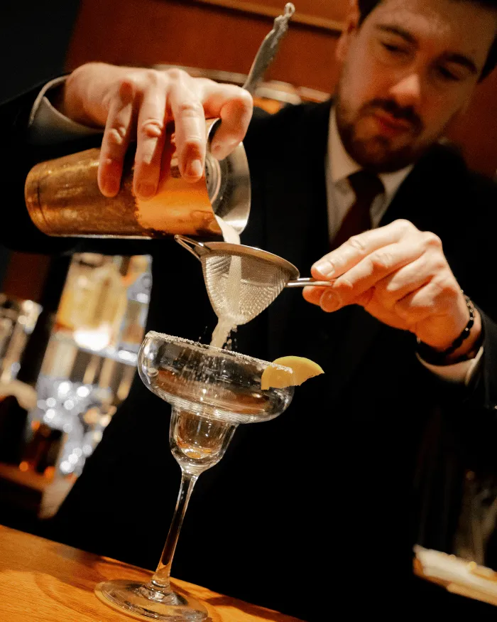 Ein Barkeeper filtert einen Cocktail in ein Margaritaglas mit einer Zitronenscheibe am Rand.