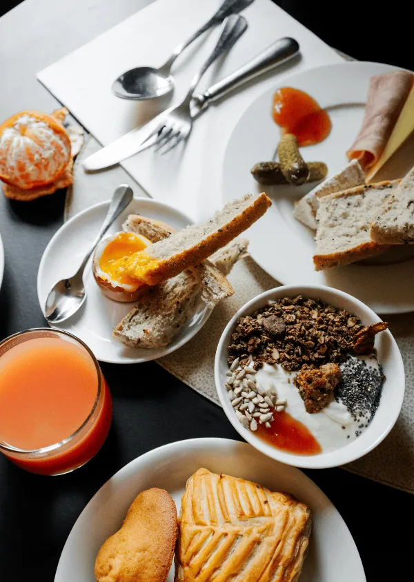 Frühstück bestehend aus Orangensaft, geschälter Mandarine, gekochtem Ei mit Toast, Joghurt mit Müsli und Samen, Brot mit Essiggurken, Schinken und Käse sowie zwei Backwaren.
