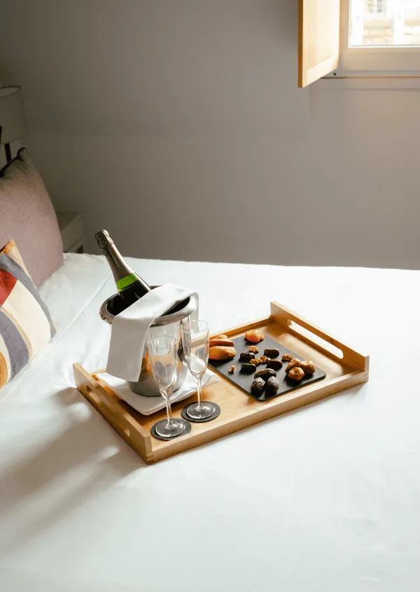 Holztablett auf einem weißen Bett mit einer Flasche Champagner in einem Eiskübel, zwei Flöten und einem Tablett mit kleinen, abwechslungsreichen Snacks.