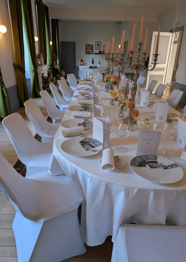 Large table set for an elegant dinner with white tablecloths, decorated plates, multi-pronged candle holders, and chairs covered with white covers in a room with large windows decorated with green curtains.
