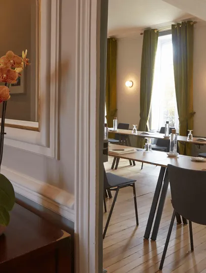 Bright dining room with wooden tables, modern chairs, large windows with green curtains and orange orchid in the foreground.
