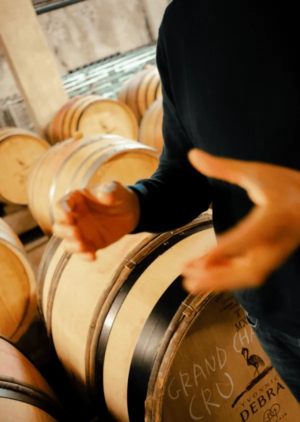 Person in a black sweater holding a glass in front of oak barrels marked 'Grand Cru' in a wine cellar.