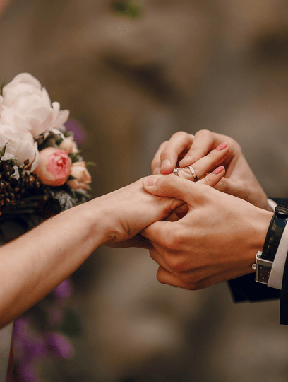 Eine Person legt während einer Zeremonie einen Verlobungsring an die linke Hand einer anderen Person, mit einem Blumenstrauß im Hintergrund.