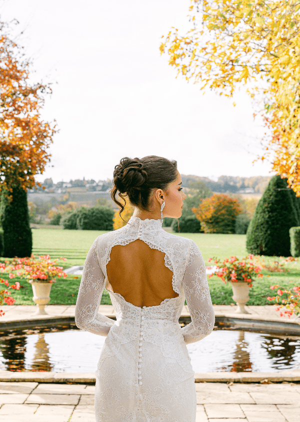 Femme vue de dos portant une robe de mariée en dentelle blanche avec un dos nu, debout devant un bassin décoré de fleurs en automne.