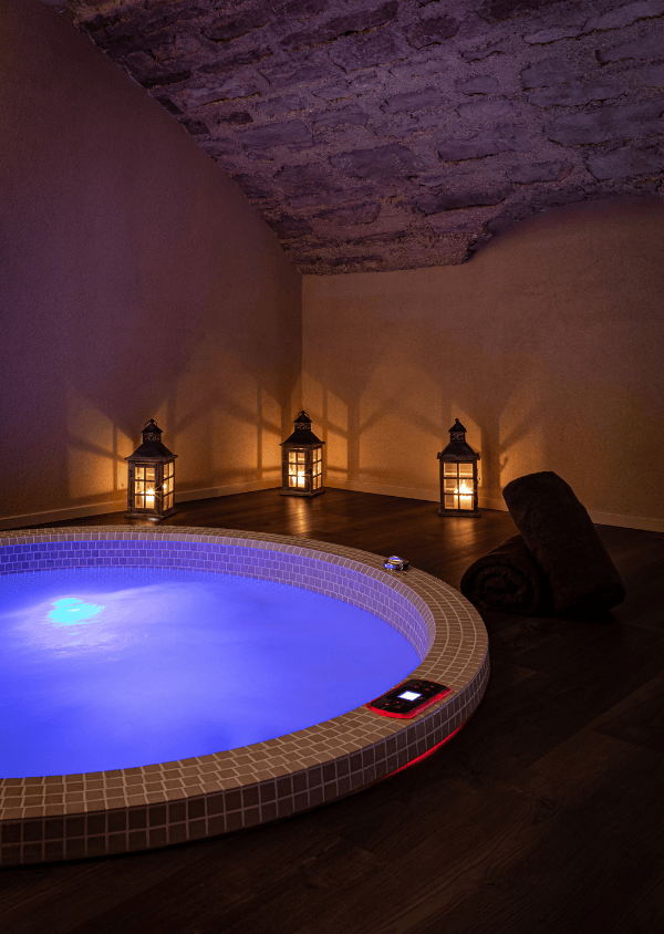 Vasca idromassaggio rotonda illuminata di luce blu con tre lanterne accese appoggiate a un muro di pietra e due asciugamani scuri arrotolati su un pavimento di legno.