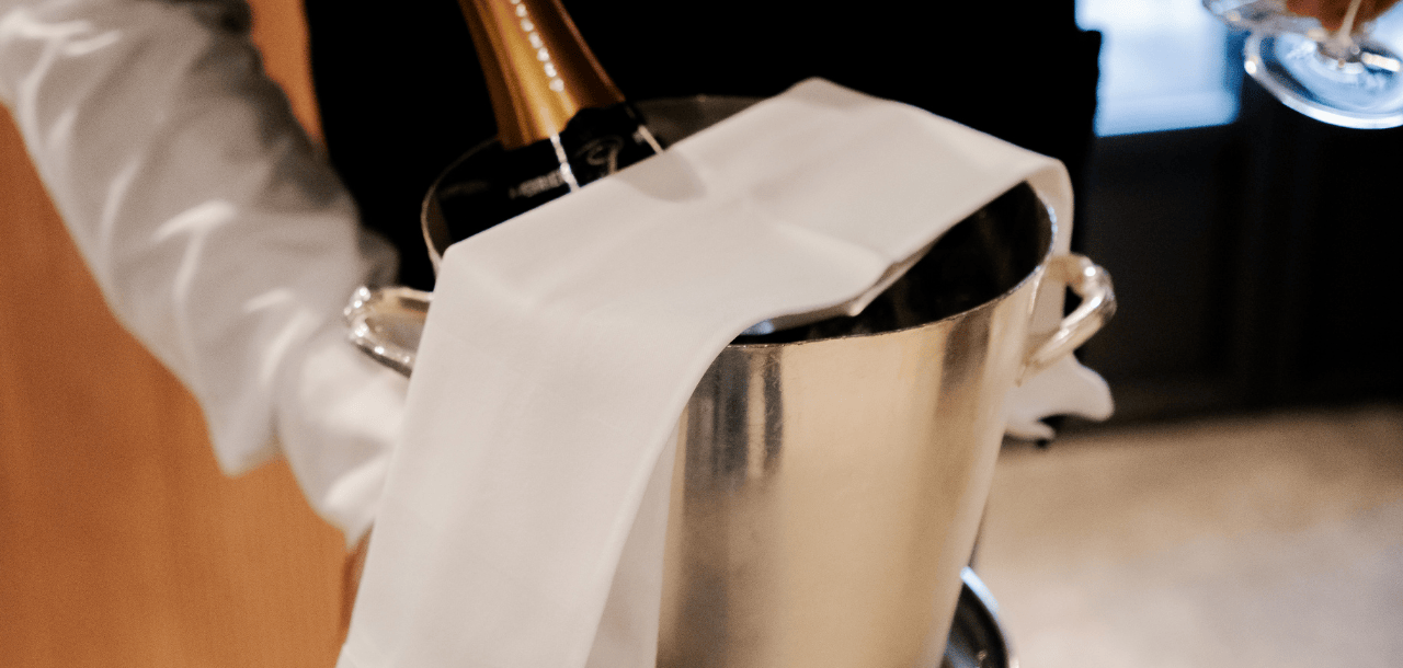 Un cameriere che trasporta un secchio per champagne in acciaio inossidabile con una bottiglia e un tovagliolo bianco in un ambiente lussuoso.