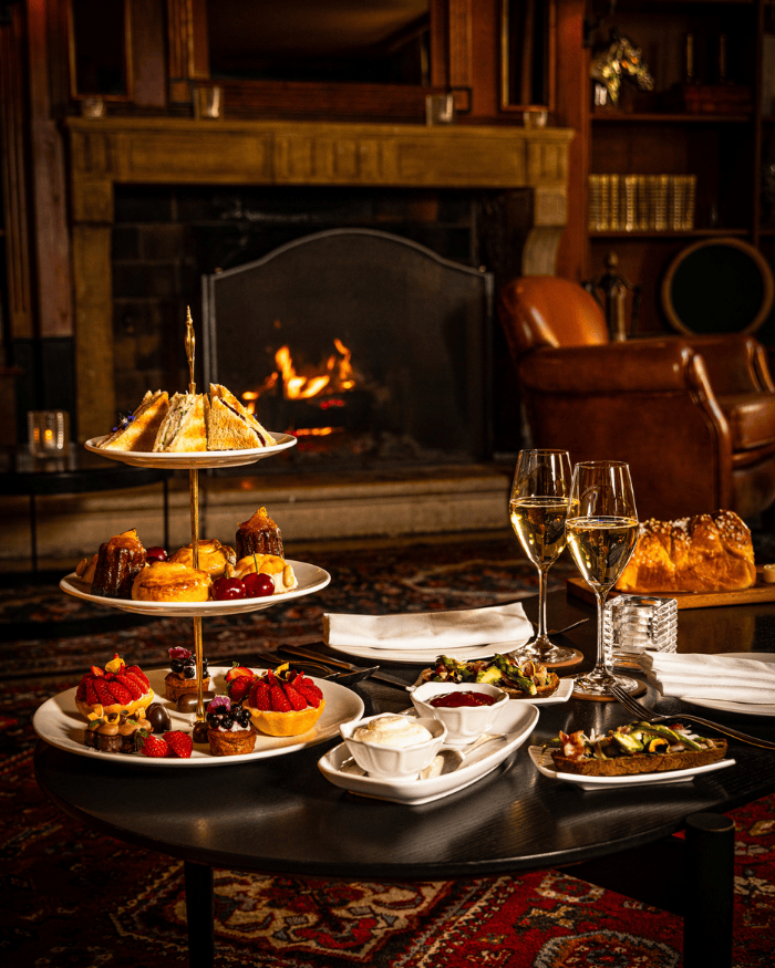 Luxuriöse Teezeit wird in einer gemütlichen Lounge mit offenem Kamin serviert, darunter ein Gebäckstand, Scones mit Sahne und Marmelade sowie zwei Gläsern Champagner.