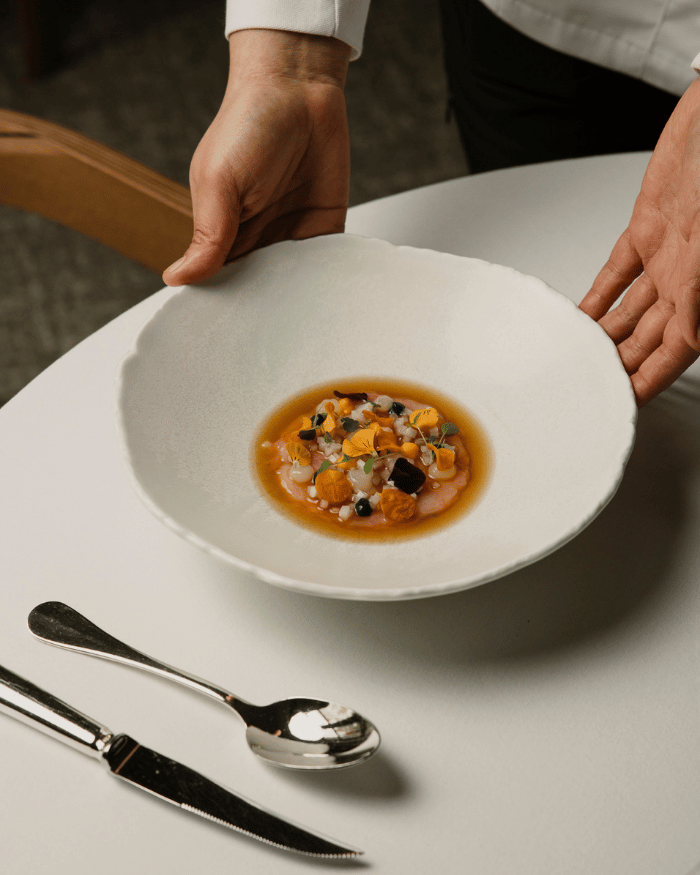 Hands serving a white bowl with a gourmet dish of seafood, sauce, and edible flowers on a white table.