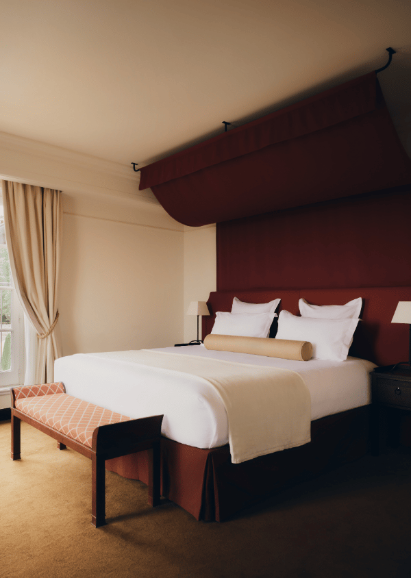Hotel room with a large bed, white linens, red headboard, bench, beige curtains, and bedside lamps.