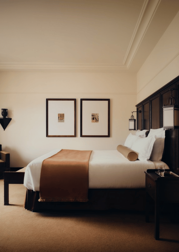Elegant hotel room with a neatly made bed, white linens, brown runner, framed artwork, and wooden furniture.