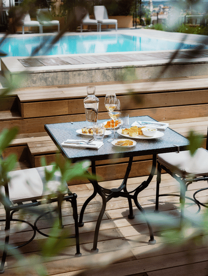 Table with glasses, carafe, and plates on a wooden deck by a pool with two chairs.