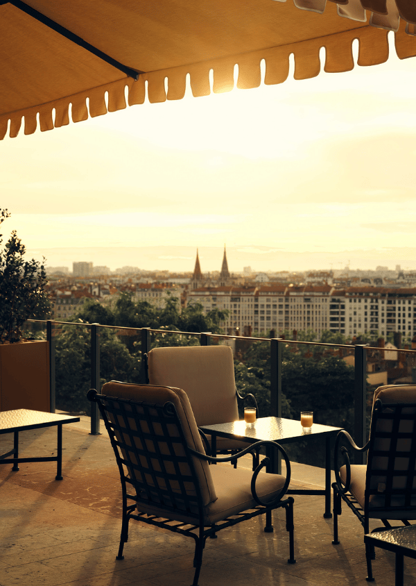 Outdoor seating area with cushioned chairs and table under awning overlooking a cityscape at sunset.