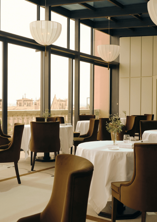 Restaurant interior with brown chairs, round tables with white tablecloths, and large windows overlooking buildings.