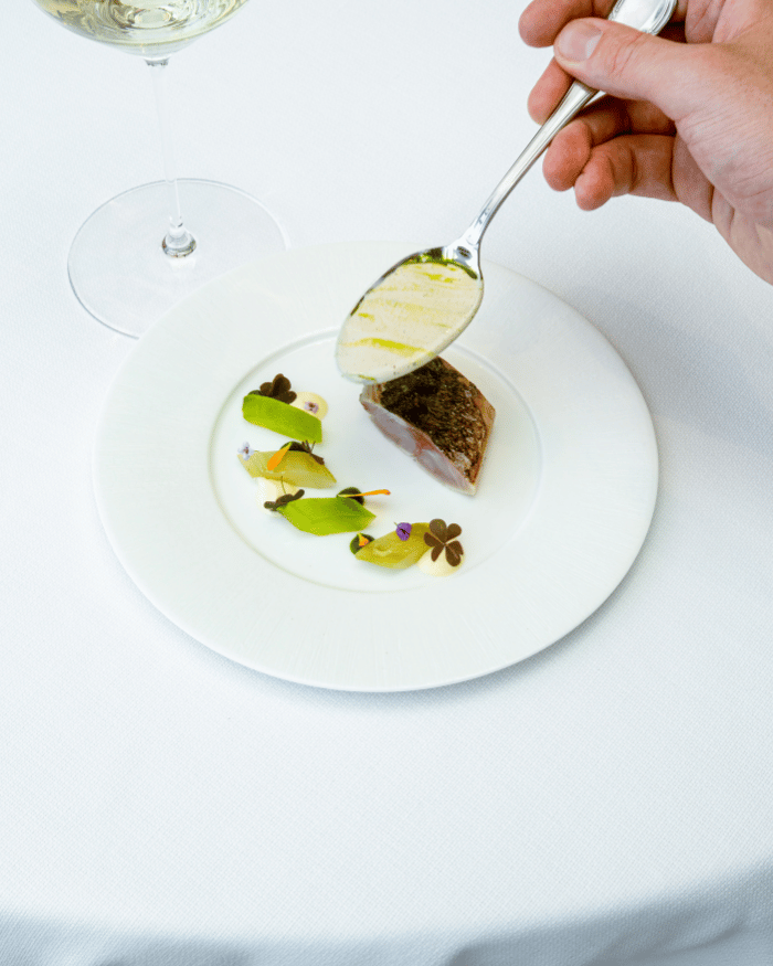 Hand with a spoon drizzling sauce over a plated gourmet fish dish with greens and white sauce dots.