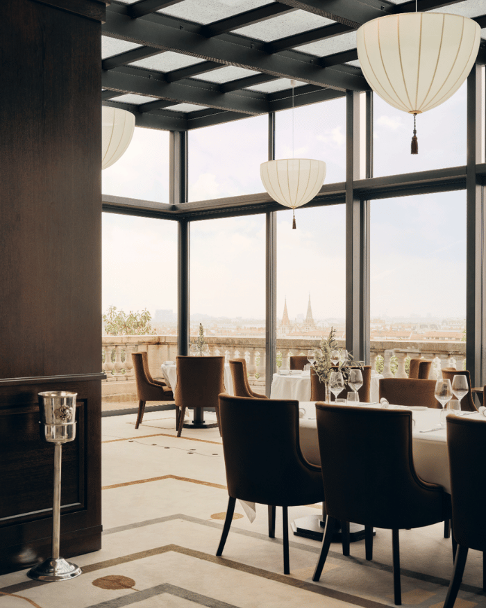 Elegant dining room with brown chairs, white tablecloths, hanging lamps, and large windows overlooking a cityscape.