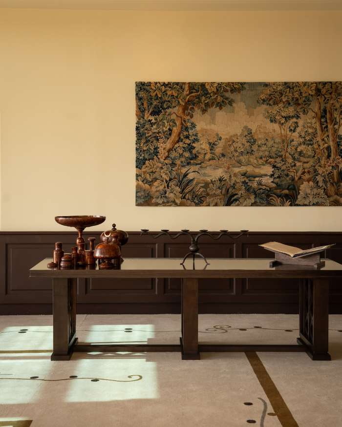 Wooden table with decorative items and an open book in a room with a tapestry on the wall.