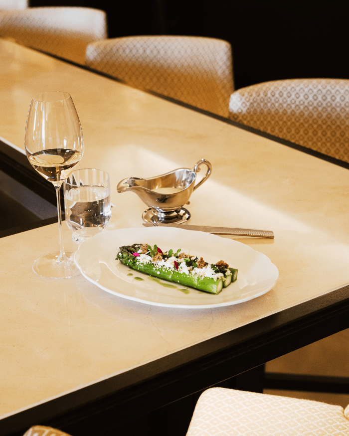 Elegant table setting with asparagus dish, glass of white wine, water glass, and sauce boat on marble surface.